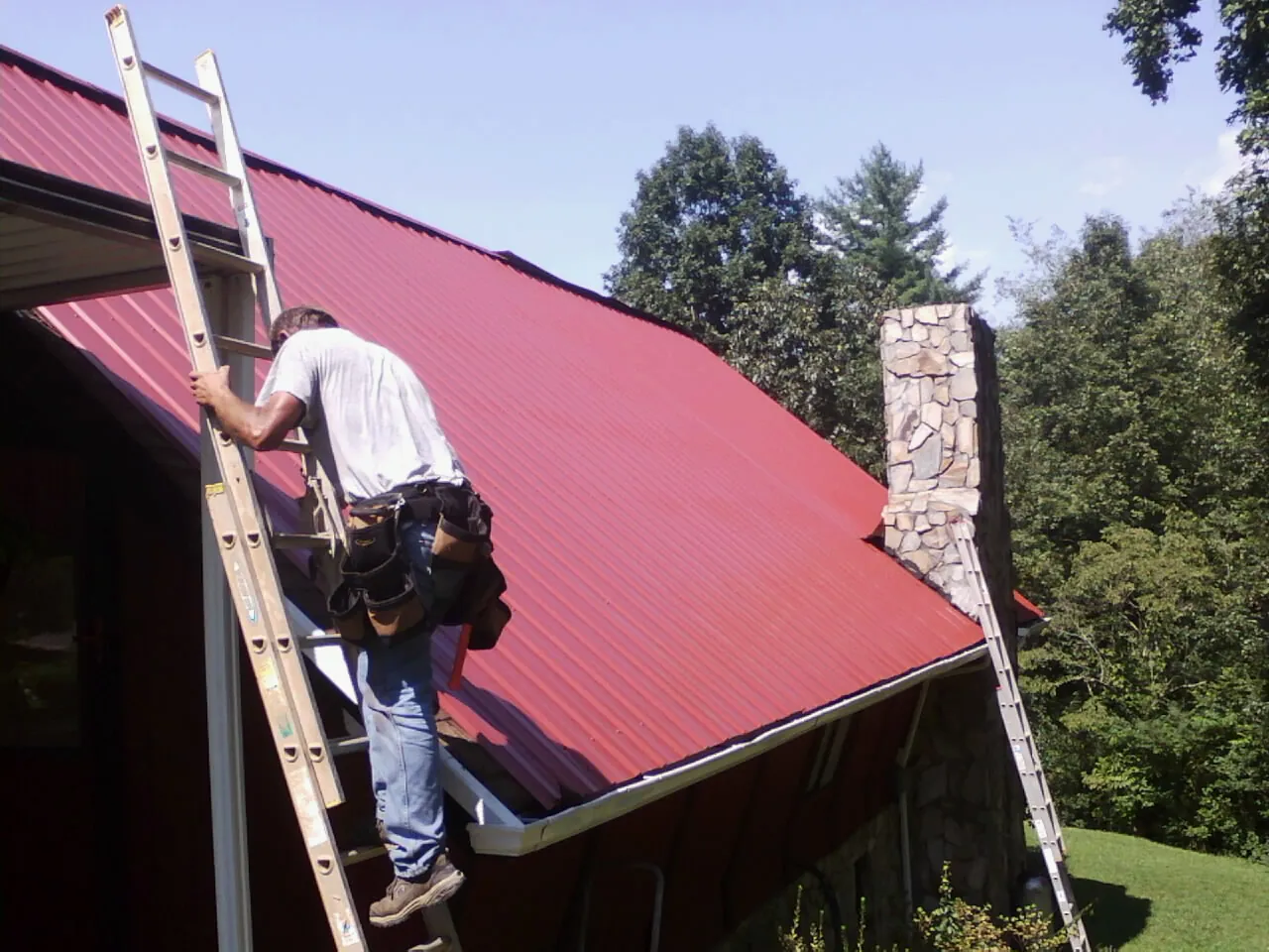 Roofer on a ladder beside a red metal roof and stone chimney — Soffit & Fascia Repair by Roofing Brothers in Blacksville